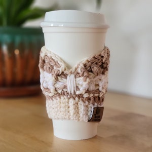 May include: A white to-go coffee cup with a crocheted beige and white cup cozy. The cozy has a bow design and a brown leather tag with the text "The Needle" on it.