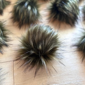 May include: Close-up of several faux fur pom-poms in shades of brown, tan, and black. The pom-poms are round and fluffy, with long, spiky strands extending outwards. The background is a light-colored wooden surface.