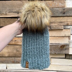 May include: A teal blue knitted beanie with a fluffy brown and black pom pom on top. The beanie has a brown leather patch on the side.
