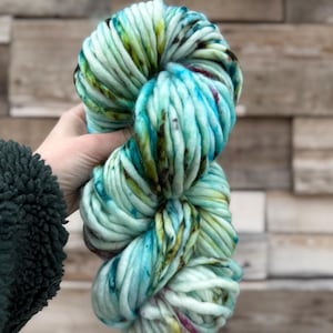May include: A large skein of chunky yarn in shades of teal, blue, green, and touches of purple. The yarn is held in a hand, showcasing its thick, textured strands. The background is a wooden wall.