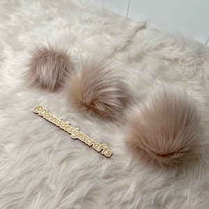 May include: Three fluffy, light brown faux fur pom poms on a white fluffy surface. The text "@Dannisdesignscrafts" is written on a wooden sign in the foreground.