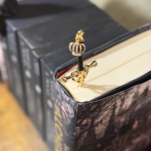 Può includere: Un segnalibro a forma di spada dorata con una corona in cima, inserito in un libro. Il segnalibro ha un'impugnatura nera ed è posto tra le pagine di un libro con una copertina scura.