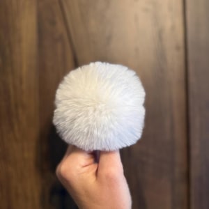 May include: A fluffy, white pom-pom, held in a hand, against a blurred wooden background. The pom-pom appears soft and round, with a dense, textured surface. The color is a bright, clean white, suggesting a clean and new item.