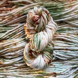 May include: A skein of thick, hand-dyed yarn featuring a variegated color palette of cream, sage green, and rust orange. The yarn is twisted into a figure-eight shape and rests atop a background of identical yarn strands, showcasing a soft texture.