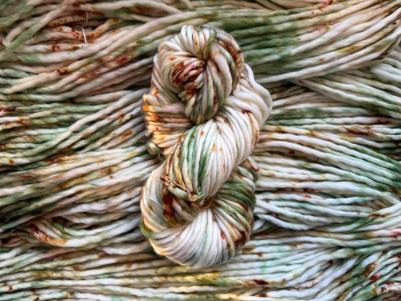 May include: A skein of thick, hand-dyed yarn featuring a variegated color palette of cream, sage green, and rust orange. The yarn is twisted into a figure-eight shape and rests atop a background of identical yarn strands, showcasing a soft texture.