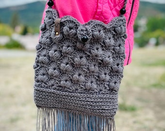 May include: A gray crocheted handbag with a fringe detail and a black button closure. The bag is worn over the shoulder of a person wearing a pink jacket and blue jeans.