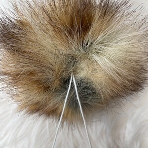 May include: A close-up of a brown and white fur pom-pom with two white strings tied around the center.