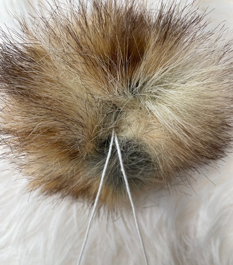 May include: A close-up of a brown and white fur pom-pom with two white strings tied around the center.