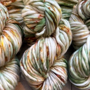May include: A collection of thick, chunky yarn skeins featuring a variegated color palette of cream, sage green, and burnt orange speckles. The yarn is twisted into soft, voluminous bundles, showcasing a multi-tonal appearance with a soft texture.