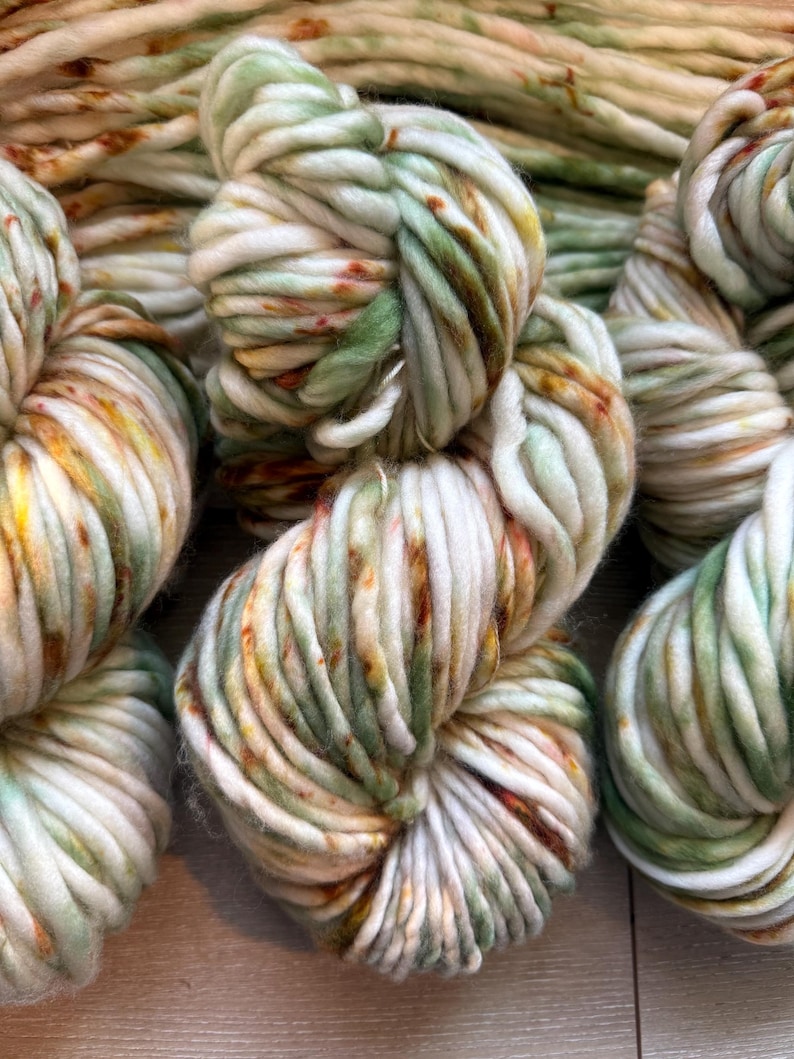 May include: A collection of thick, chunky yarn skeins featuring a variegated color palette of cream, sage green, and burnt orange speckles. The yarn is twisted into soft, voluminous bundles, showcasing a multi-tonal appearance with a soft texture.