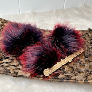 May include: Three fluffy faux fur pom poms in shades of black and red, resting on a woven basket. The pom poms have a textured appearance, with the red streaks adding visual interest. The basket is made of natural-colored woven material.
