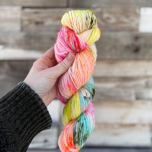 Puede incluir: Un ovillo de hilo teñido a mano en una vibrante gama de colores. El hilo transita del crema al rosa brillante, naranja, amarillo y turquesa, con acentos en verde oscuro y negro. El hilo se sostiene en una mano sobre un fondo de madera.