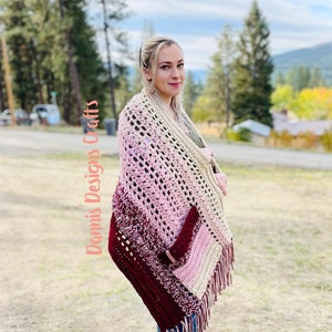 May include: A woman wearing a crocheted shawl with a gradient of pink, beige, and burgundy colors. The shawl has a fringe border and a pocket on the side. The text "Damnis Designs Crafts" is visible on the left side of the image.