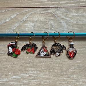 May include: Six gold-tone metal stitch markers with black and red enamel designs. The designs include a skull with roses, a bat with a crescent moon, a potion bottle with a heart, a bat with a skull, and two bats with roses.