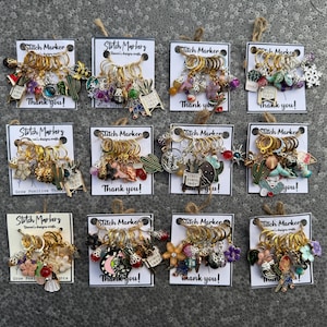 May include: A collection of gold-toned stitch markers, each adorned with various charms, including cacti, flowers, and miniature objects. Each set is attached to a card with the text "Stitch Marker" and "Thank you!"