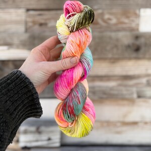 Puede incluir: Un ovillo de lana colorida en tonos rosa, amarillo, naranja y turquesa. La lana se sostiene en una mano, sobre un fondo de madera. La lana parece suave y es adecuada para proyectos de tejido o ganchillo.