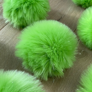 May include: Several bright green fluffy pom-poms with silver glitter accents are scattered across a wooden surface. The pom-poms are round and have a soft, textured appearance. The background is a light brown wood grain.