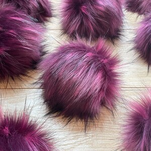 May include: Close-up of several fluffy, round pom-poms. The faux fur is a deep burgundy color with streaks of pink and black. The pom-poms are arranged on a light-colored wooden surface, creating a textured and colorful display.