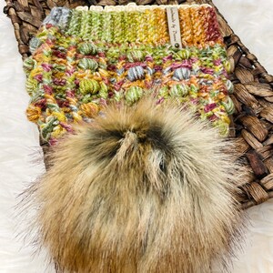 May include: A crocheted hat with a pom pom made of faux fur. The hat is a mix of green, yellow, orange, and brown colors. The hat has a label that says "Handmade by Dream".