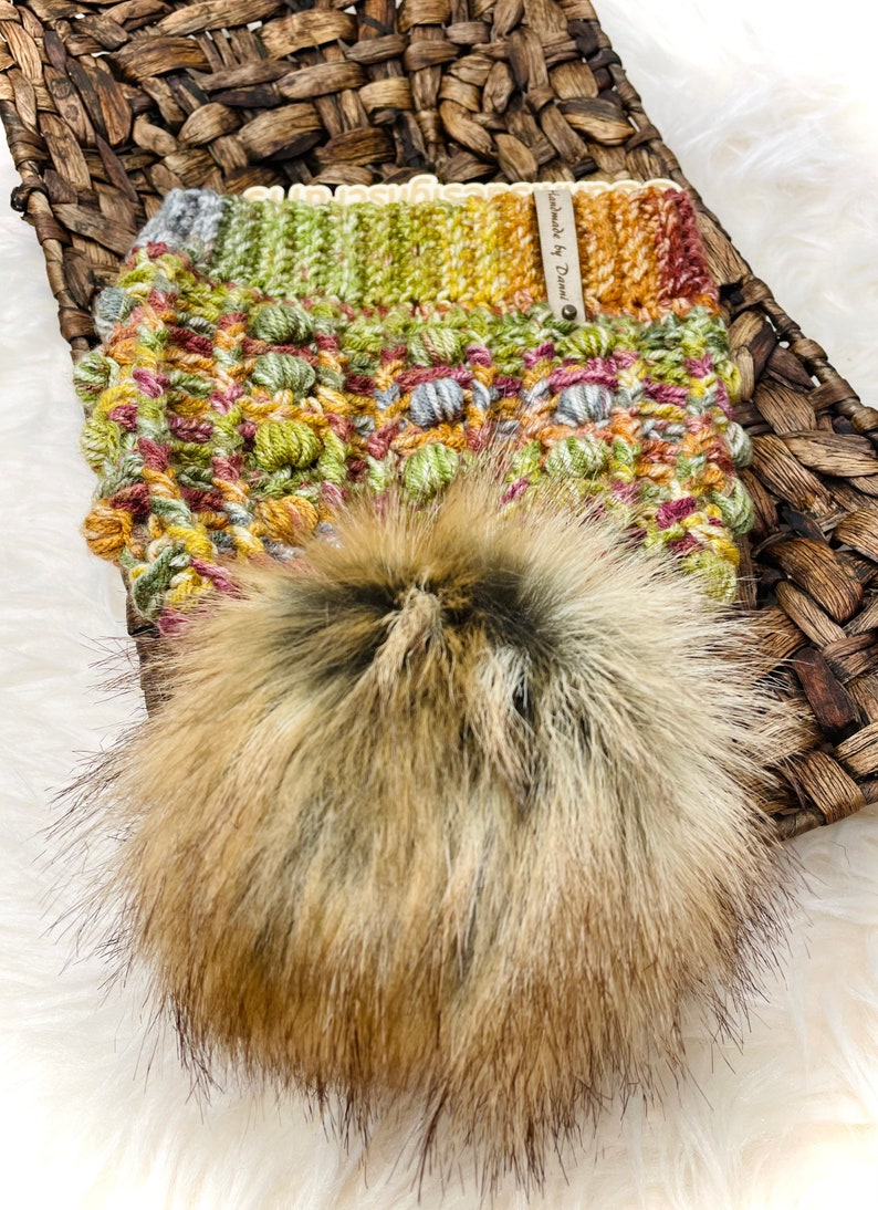 May include: A crocheted hat with a pom pom made of faux fur. The hat is a mix of green, yellow, orange, and brown colors. The hat has a label that says "Handmade by Dream".