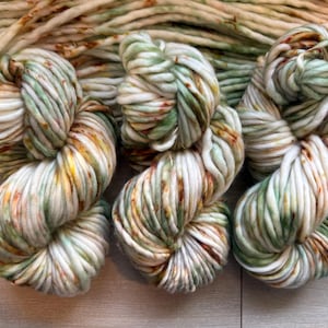 May include: Three skeins of thick, chunky yarn featuring a variegated color palette of cream, sage green, and burnt orange speckles. The yarn is twisted into dense, rounded bundles, showcasing a soft, bulky texture with consistent fiber thickness.