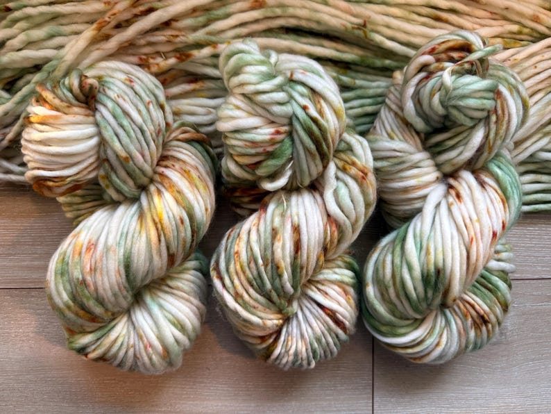 May include: Three skeins of thick, chunky yarn featuring a variegated color palette of cream, sage green, and burnt orange speckles. The yarn is twisted into dense, rounded bundles, showcasing a soft, bulky texture with consistent fiber thickness.
