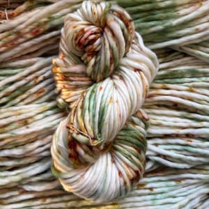 May include: A thick, hand-dyed skein of yarn featuring a creamy off-white base color with variegated splashes of sage green and burnt orange throughout. The yarn has a twisted, multi-ply texture and is displayed against a background of itself.