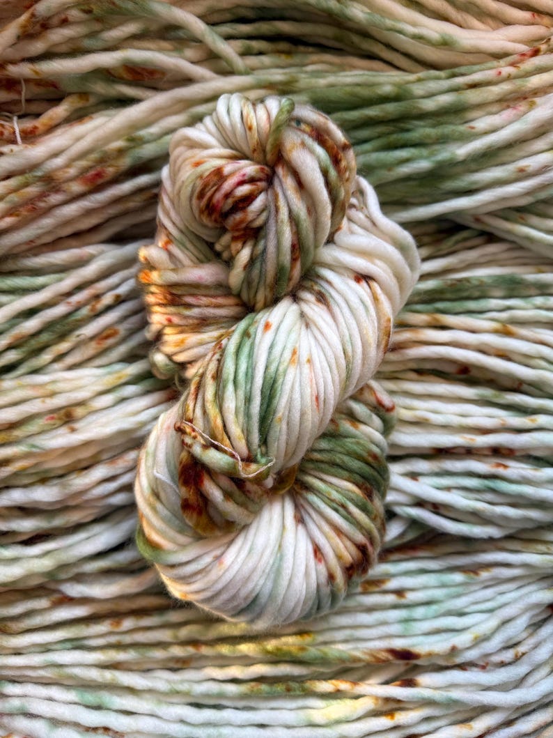 May include: A thick, hand-dyed skein of yarn featuring a creamy off-white base color with variegated splashes of sage green and burnt orange throughout. The yarn has a twisted, multi-ply texture and is displayed against a background of itself.