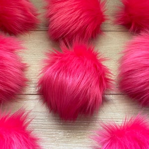 May include: A collection of ten bright pink fluffy pom poms. The pom poms are arranged in a symmetrical pattern on a light brown wooden surface.