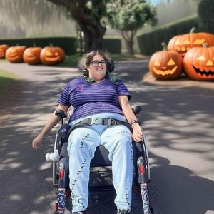 May include: A person in a wheelchair wearing a purple and white striped shirt and blue jeans with stitched details is smiling at the camera. The person is in front of a row of jack-o'-lantern pumpkins.