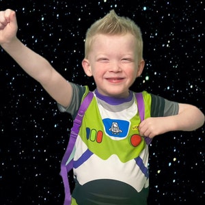 May include: A young boy wearing a green, white, and purple Buzz Lightyear costume with a purple backpack. He is smiling and has his arm raised in the air. The background is a black starry sky.