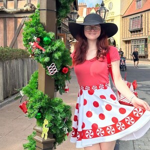 Puede incluir: Una mujer con una camisa roja y una falda blanca con diseños de bastones de caramelo rojos y blancos. Lleva un sombrero negro y gafas. Está de pie frente a un edificio decorado con luces navideñas.