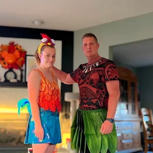May include: A woman and a man wearing colorful costumes. The woman is wearing a red, orange, and yellow top with a blue skirt and a rooster headpiece. The man is wearing a red, black, and white patterned shirt with a green grass skirt.