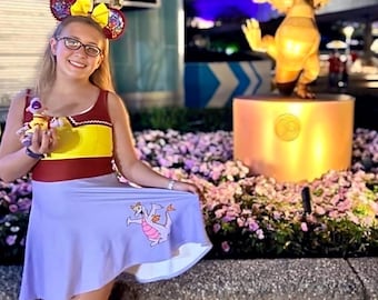 Puede incluir: Un vestido sin mangas con cuerpo amarillo, ribete granate y falda de color violeta claro. La falda muestra un pequeño gráfico de un dragón de color violeta y rosa. El conjunto se completa con orejas de ratón de lentejuelas y zapatillas blancas.