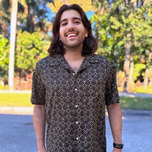 Pode incluir: Um homem vestindo uma camisa de botões de manga curta preta e dourada com um padrão geométrico.