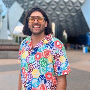 May include: A man wearing a white button-down shirt with a colorful pattern of various Disney park logos. He is smiling and wearing sunglasses. The background is a large, gray, geodesic dome.