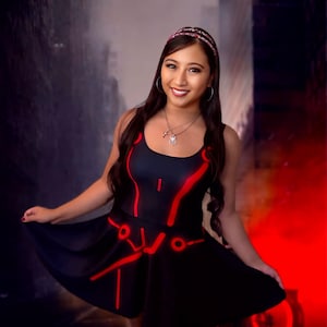May include: Black skater dress with red neon-style detailing. The dress has a scoop neckline and a flared skirt. The design incorporates lines and circles, giving a futuristic look. The dress is sleeveless and made of a smooth material.