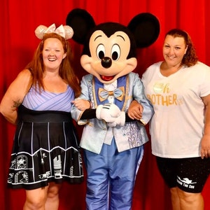 May include: Mickey Mouse, dressed in a blue suit with a gold bow tie, poses with two people. The person on the left is wearing a purple and black dress with a white bow in her hair. The person on the right is wearing a white t-shirt with the text "Oh Brother" and black shorts with the text "Under The Sea".