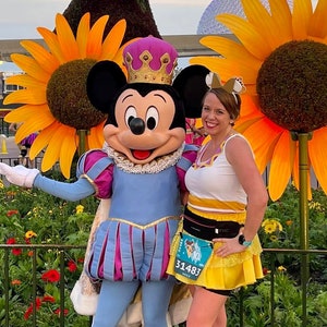 Puede incluir: Mickey Mouse vestido de príncipe, con un traje azul y morado con una corona, posa con una mujer que lleva una falda amarilla y una blusa blanca. La mujer sostiene un dorsal de carrera con el número 31483. El fondo presenta un topiario de Goofy y girasoles.