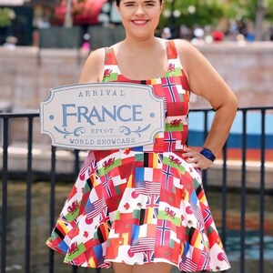 Puede incluir: Un vestido sin mangas con un estampado de varias banderas internacionales, incluyendo las de Estados Unidos y Gales. El vestido se muestra junto a un letrero de color claro que dice Arrival France Epcot World Showcase.