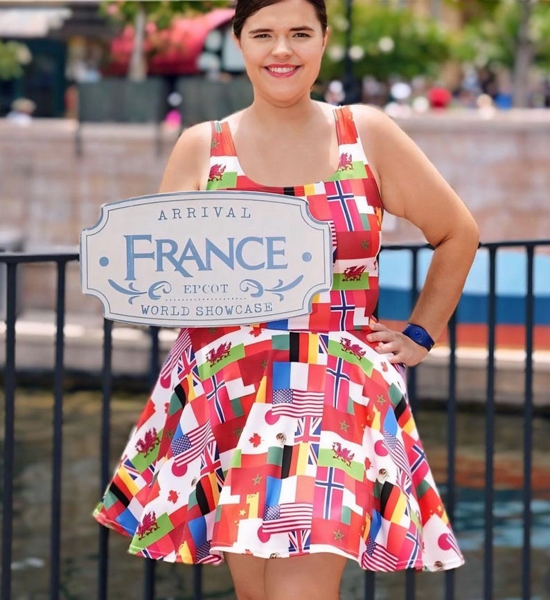 Puede incluir: Un vestido sin mangas con un estampado de varias banderas internacionales, incluyendo las de Estados Unidos y Gales. El vestido se muestra junto a un letrero de color claro que dice Arrival France Epcot World Showcase.