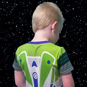 May include: A young child wearing a green, white, and blue t-shirt with a cartoon spaceship design on the back. The child is facing away from the camera and looking down. The background is a black starry sky.