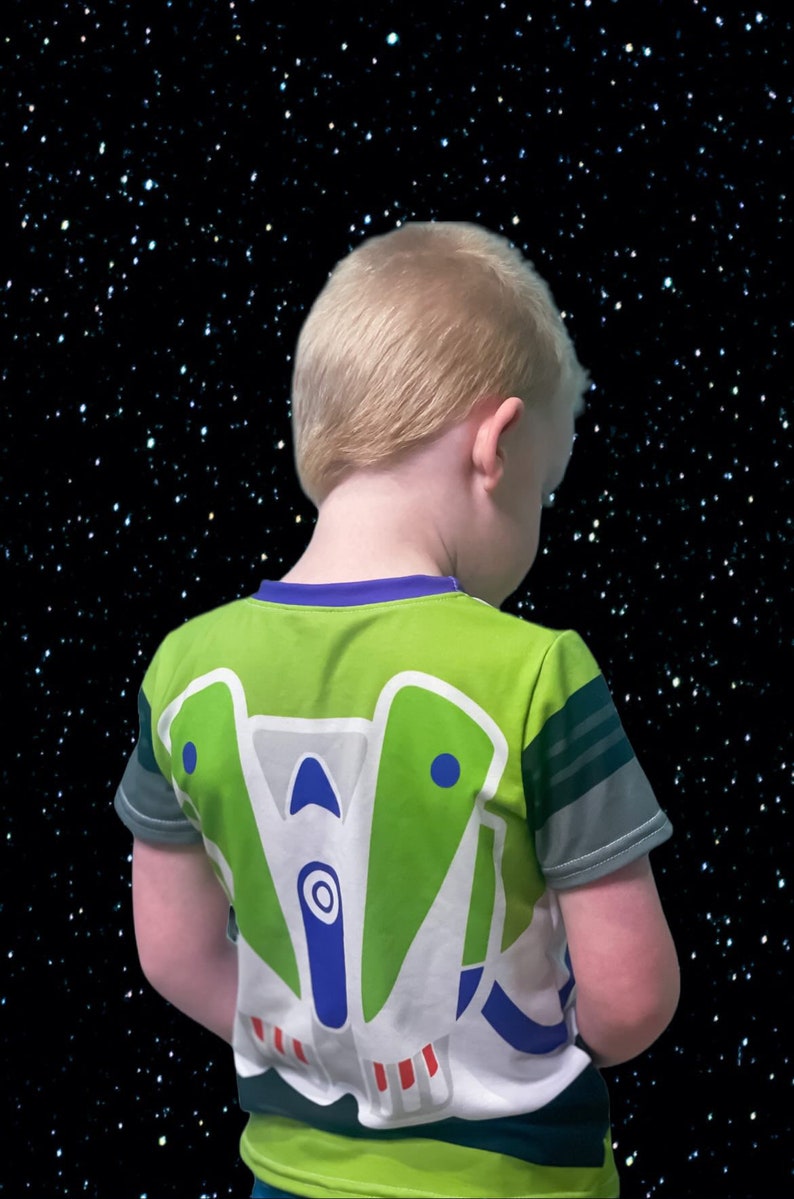 May include: A young child wearing a green, white, and blue t-shirt with a cartoon spaceship design on the back. The child is facing away from the camera and looking down. The background is a black starry sky.