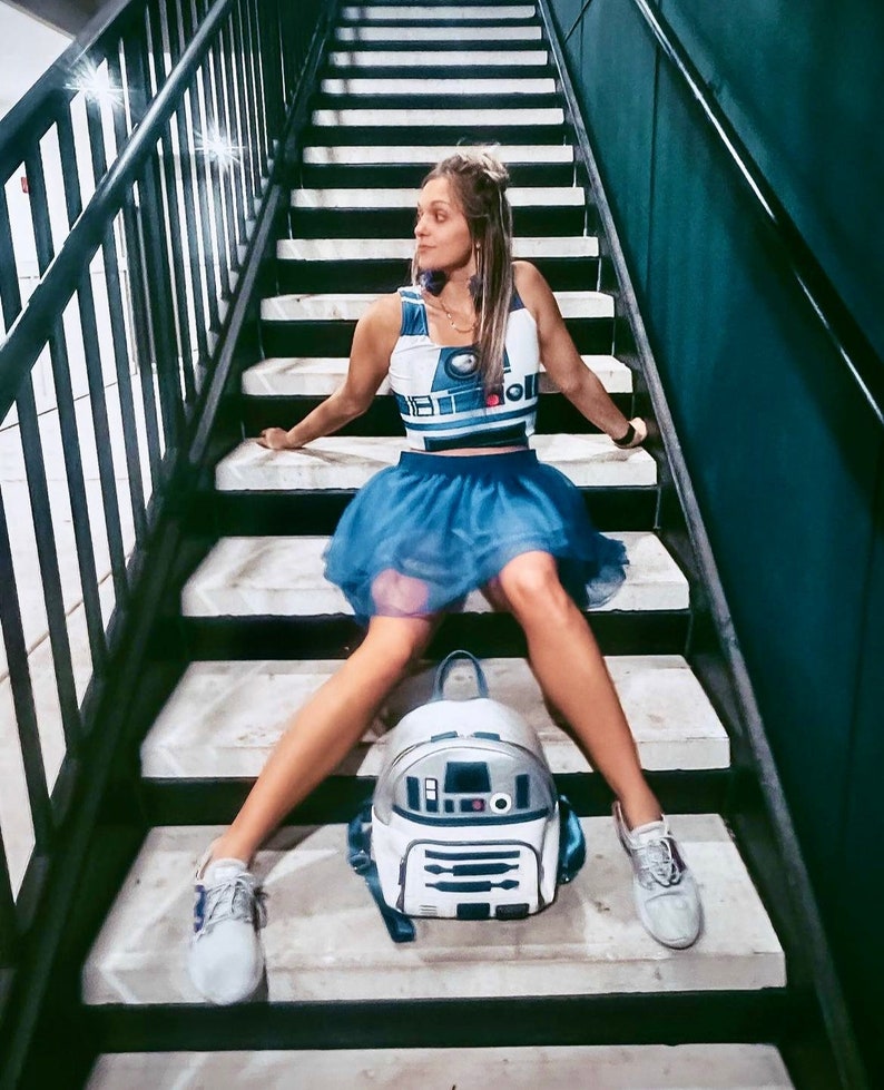 May include: A woman wearing a blue skirt and a white crop top with a Star Wars R2-D2 design sits on a set of stairs. She is wearing white trainers and has a white R2-D2 backpack on her back.