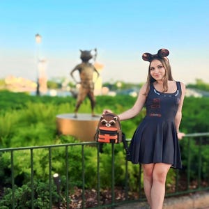 May include: A woman in a dark blue dress and mouse ear headband, holding a Rocket Raccoon backpack. A bronze Rocket Raccoon statue is in the background. The dress has a flared skirt and the backpack has orange detailing.