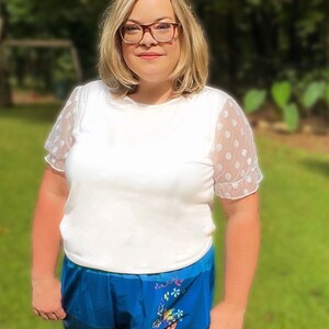 May include: A woman wearing a white shirt with polka dot sleeves and blue shorts with a floral print.
