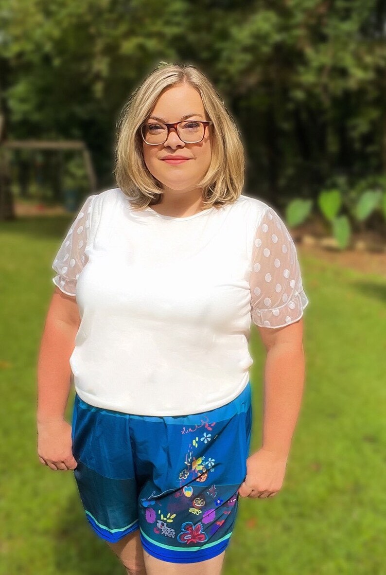 May include: A woman wearing a white shirt with polka dot sleeves and blue shorts with a floral print.