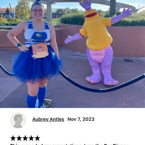 May include: A woman wearing a blue and white crop top with a blue tutu skirt and blue socks poses with a purple character dressed in a yellow shirt. The woman is holding a race bib that says "Wine & Dine Half Marathon".