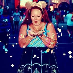 May include: A woman wearing a blue and white striped tank top and a black skirt with a white design blows confetti into the air. She is wearing a gold Mickey Mouse ear headband with a pink bow.