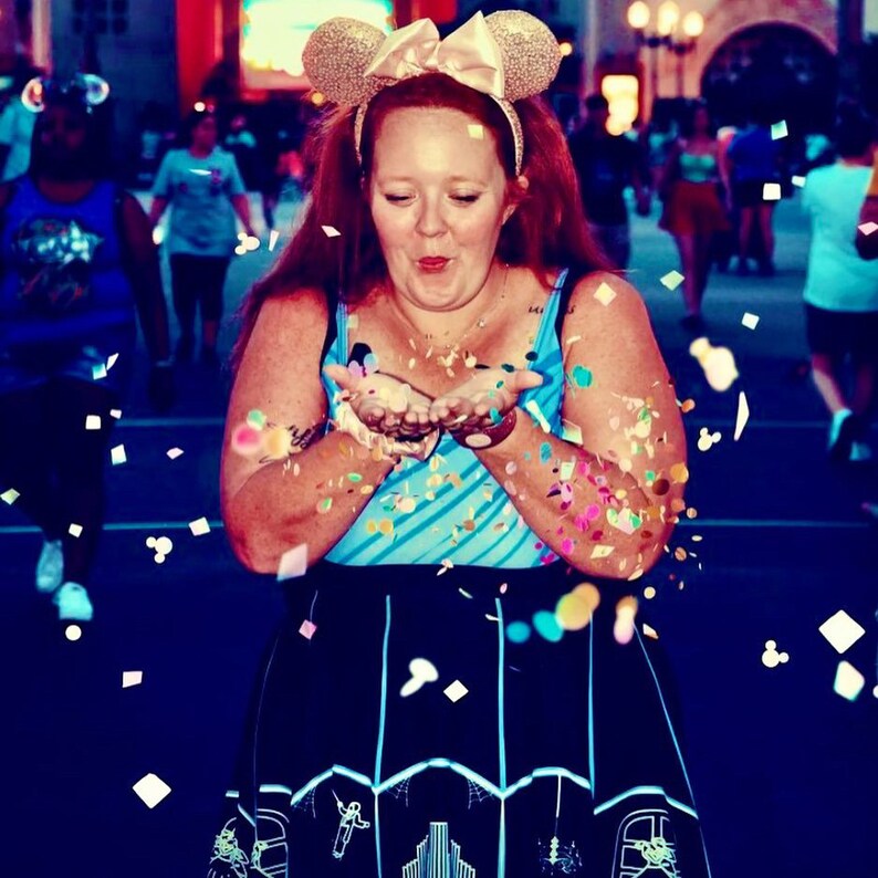 May include: A woman wearing a blue and white striped tank top and a black skirt with a white design blows confetti into the air. She is wearing a gold Mickey Mouse ear headband with a pink bow.
